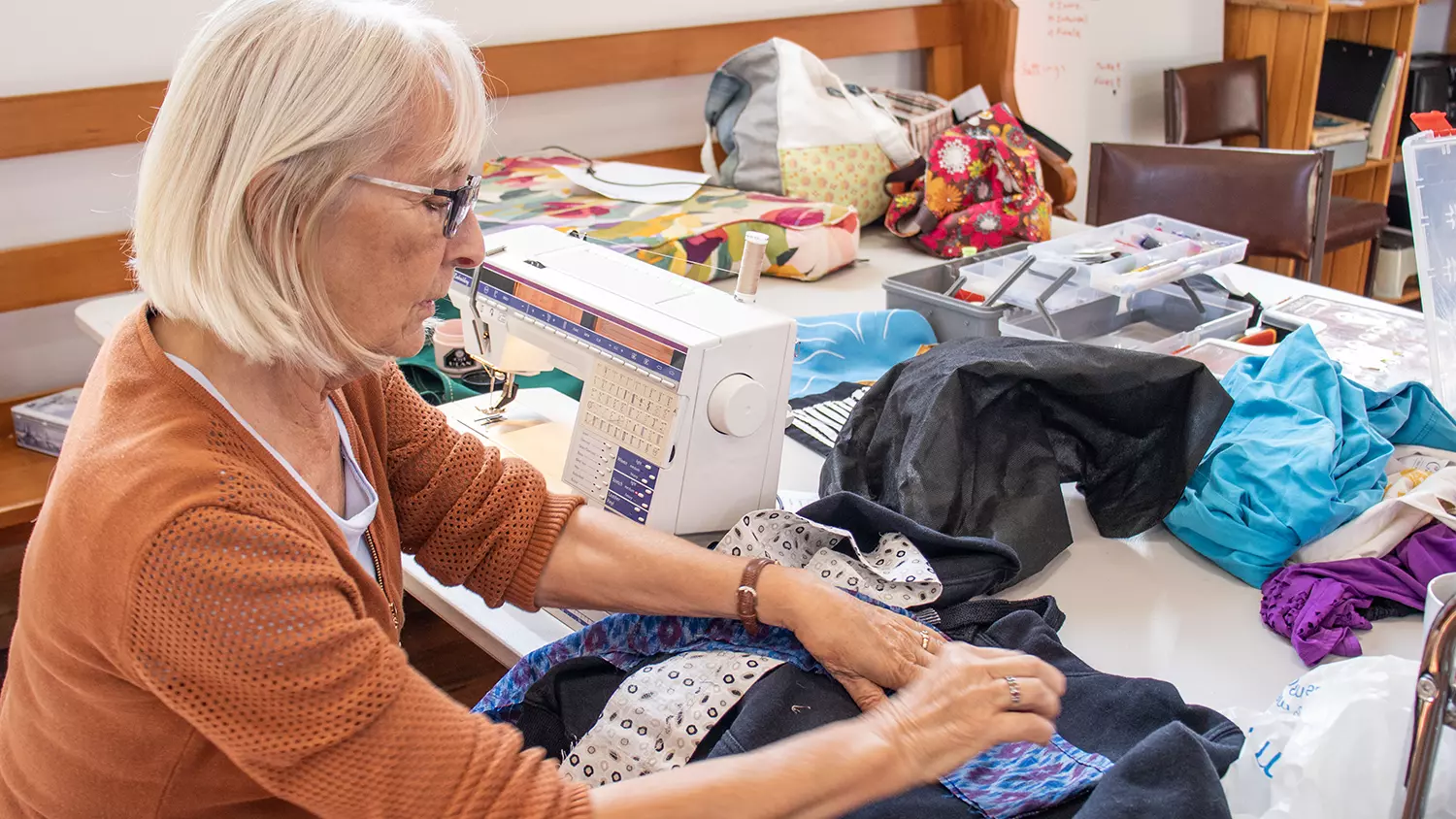 A volunteer at the SHASA Repair Café sits at a large table with her sewing machine, surrounded by fabric and sewing equipment, working on patching an item of clothing.